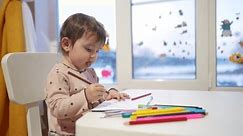 赤ちゃんの女の子は、自宅で窓のそばのテーブルに座っている間に描きます。 ハッピーファミリーキッドコンセプト。赤ちゃんの娘は、室内で紙のシートに鉛筆で描くことを学びます。夢の細かい運動能力の開発
