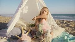 Funny little blonde girl pointing her finger while singing expressively song out loud in tent on beach. Emotional little girl with long hair flying at wind. Playful child funny face at ocean at sunset