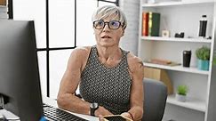 A thoughtful mature woman with glasses and short grey hair using a smartphone in an office setting.