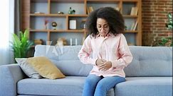 Upset young african american female suffering from abdominal pain sitting on sofa in room at home. Black woman keeps her hands on her stomach. She has poisoning, constipation, bloating or menstruation