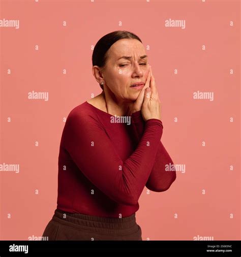 Elderly woman in red presses her cheek with both hands, showing toothache or facial pain ...