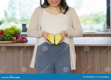 Asian Pregnant Woman Holding Fresh Avocado Fruit Close To Her Belly in Kitchen at Home. Healthy ...