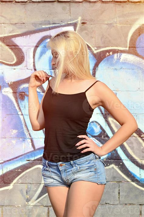 Sexy caucasian blonde girl in denim shorts and black tank top posing against graffiti wall in ...