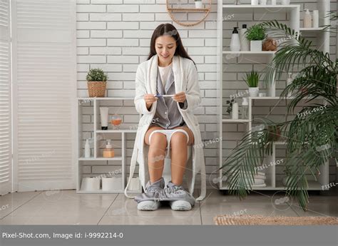 Happy young Asian woman with pregnancy test sitting on toilet bowl in ...