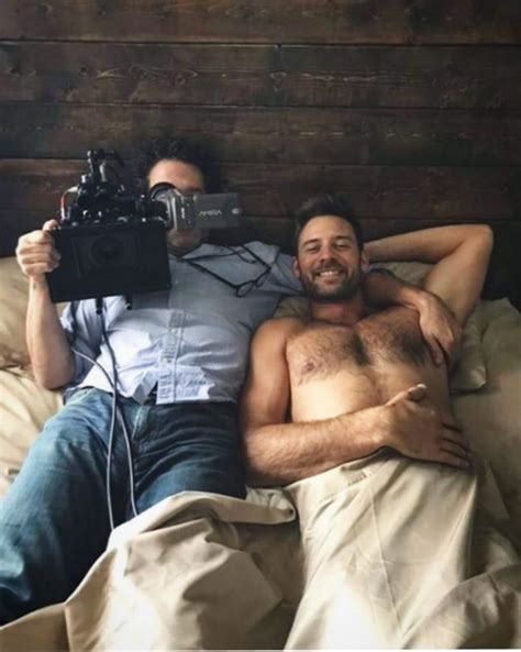 two men laying in bed and one is holding a camera