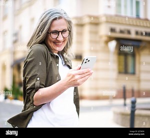 Mature beautiful grey hair woman looking at smartphone walking at the streets of old European town. Senior businesswoman read text message, answering video call standing outdoors. Selective focus Stock Photo - Alamy