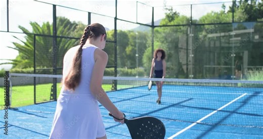 Diverse women playing padel, partner tossing ball, white-dress woman returning with racket for fun