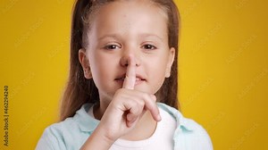 Keep Silence. Funny little girl showing shh sign, holding finger on lips and looking at camera, orange studio background
