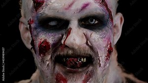 Close-up face of zombie man with makeup with fake wounds scars and white contact lenses looking at camera and clicks his teeth, trying to scare in dark room. Sinister dead guy. Halloween, filming
