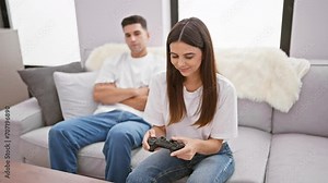 In a cozy living room, a woman plays video games while a man watches from the couch, highlighting leisure and couple dynamics indoors.