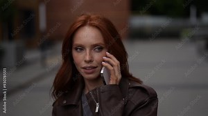 Charming young woman with long red hair and freckles smiling while chatting on smartphone outdoors. Pretty redhead female dressed in brown leather jacket, enjoying conversation in urban setting