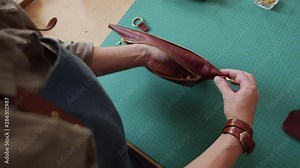 Female Caucasian tanner wearing apron standing at worktop in tannery and attaching fasten rings to small leather bag