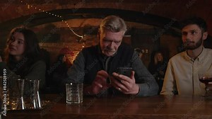 Three people drinks at the bar counter. Adult man checking his smartphone when drinking alcohol on pub terrace. Old man watching funny video using phone inside cafe. Three strangers chilling after