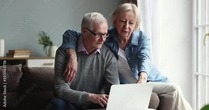 Vidéo Stock Positive senior couple in love using laptop together, sitting on home couch, hugging, shopping on Internet, pointing at computer monitor, display, smiling laughing