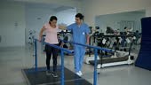 Young female patient walking with effort leaning on parallel bars...