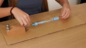 Hands pull a spring scale to measure friction on a cardboard surface using weights, highlighting physics principles