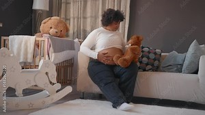 African american woman sitting alone in cozy apartment, stroking pregnant belly. Lady looking at teddy bear, waiting for her first baby, eighth month of pregnancy, firstborn in family, motherhood Stock Video