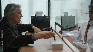 Male bank employee showing elderly woman where to sign her documents across glass teller window in office