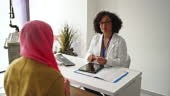 A Muslim woman sits across from her female doctor as she talks with...