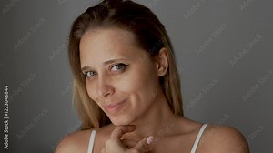 Young smiling blonde woman showing the result of plastic surgery buccal fat removal running her hand over the cheek on a dark grey background. A female face with clear highlighted cheekbones