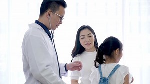 小児科医と子供のコンセプト。 アジアの男性医師は、女性の子ども患者の背中に聴診器を付け、女の子が息を吸い込んだり吐いたりする間に機能する肺の音を聞き、その上に立つ母親も耳を傾けた。