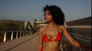 Curly-haired woman in red bikini walking on a bridge, gazing at the sunset