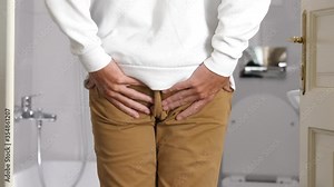 Thin man enters bathroom and clutches his buttocks because of sudden severe pain in anus area, massages this area. Medium shot, real time