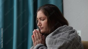 Adult woman covered with warm blanket feels unwell in cold house. Brunette housewife sneezes sitting with tired and exhausted expression in apartment