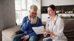 An elderly man with gray hair and a lush beard puts on glasses to read what a brunette woman doctor in a white coat wants to convey to him during a home examination on the sofa in a modern apartment | Premium Stock Video Footage