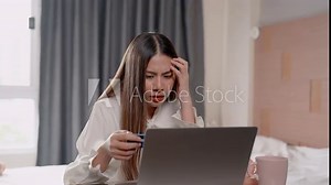 Asian girl sitting in bedroom, ordering online on laptop placed in front, lowered head pick up credit card that was on table in order pay, puzzled expression on face as credit card couldn be used.
