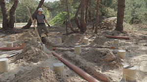 A worker, clad in work attire and equipped with safety gear, is in the process of laying large underground pipes among the partially buried concrete foundations in a forested construction site.