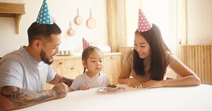 Asian girl kid wearing pink cone cap celebrates birthday with happy parents. Excited daughter sitting at table makes wish and blows candle on cupcake