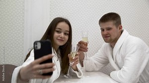 A young couple in white terry robes talking in the spa and drinking champagne. The girl takes a selfie on her smartphone. Vacation