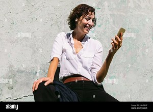 Delighted female in white formal shirt and pants sitting on concrete border and having video call Stock Photo - Alamy