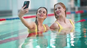 Young females in the swimming pool, two beautiful girls in yellow swimsuits take a selfie on a smartphone, girls smile and pose for the camera.