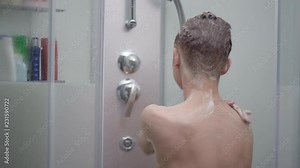 Young boy bathing under a shower at home - back view. Beautiful teen boy taking shower and washing in the bathroom. Happy child washing head, face and body with water.