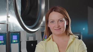 Portrait of work woman posing near industrial washing machine inside laundromat, confident employee in yellow shirt looking aside, stainless drum and control screens in background | Премиум Стоковые видеоматериалы