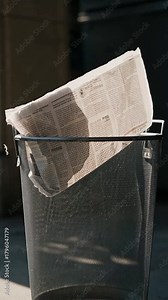 Crumpled newspaper and paper waste sticking out of a wire mesh metal trash bin in bright sunlight casting a long shadow