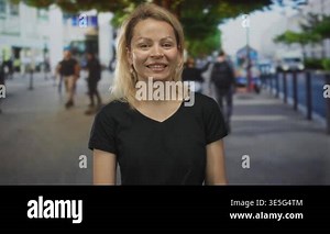 Woman pointing finger to camera with smiling face in urban street setting with blurred pedestrians; invitation connection warmth.