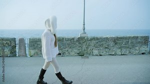 Woman walks on embankment with stone fence against lake Garda on cloudy autumn day. Female tourist in white coat enjoys weekend in Italy