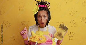 Embarrassed shocked untidy ethnic woman with combed dreadlocks says wow looks with indignant expression notices messy room poses near basket full of dirty linen holds sponge for removing dirt