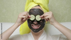 Girl in purifying black mask on face removes slices cucumber from eyes, covering it again and smiled. Charcoal face mask or black clay mud. Natural cosmetic concept. Skin care, cleansing skin.