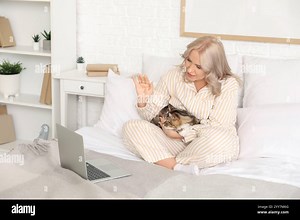 Beautiful mature woman in pajamas with cute cat and laptop video chatting on bed at home Stock Photo - Alamy