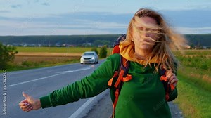 Beautiful blond girl with backpack hitching ride. Young woman hitchhiker steps back slowly along roadside, waits for passing car to stop. Hitchhiking in countryside. Hiker, tourist, traveller