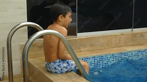 A small young boy sitting wet outside a swimming pool shivering from the cold one feels after getting out of the water.
