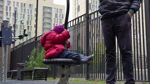 Child contemplatively views urban skyline from spinner, then begins to spin energetically, becoming vibrant blur of joyful motion, all while being watched by adult in modern city playground. Stock Video