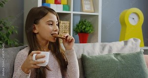 Pretty brunnette schoolgirl eating a chocolate and drinking a hot tea while sitting on the sofa and thinking about something in the nice living room. Portrait shot. Indoor
