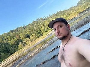 Mentally, I’m here. 🥲 Image Description: A shirtless man with a black baseball cap, a silver chain link necklace, and mask lines on his face is standing in a river with lots of trees and blue skies in the background. | Matt McGorry