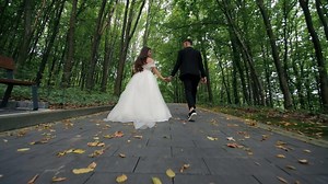 Bride and groom walking hand in hand on forest path with fallen leaves and tall trees, Romantic wedding moment in nature captured from low angle with soft natural ambiance