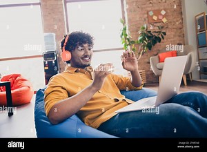 Photo portrait of attractive young man wear yellow shirt sit blue bean bag talk video call headphones modern workplace room home design Stock Photo - Alamy